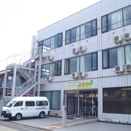 介護施設の建物外観。白いタイル張りの3階建て施設で、エントランス前に送迎車が停車している。
